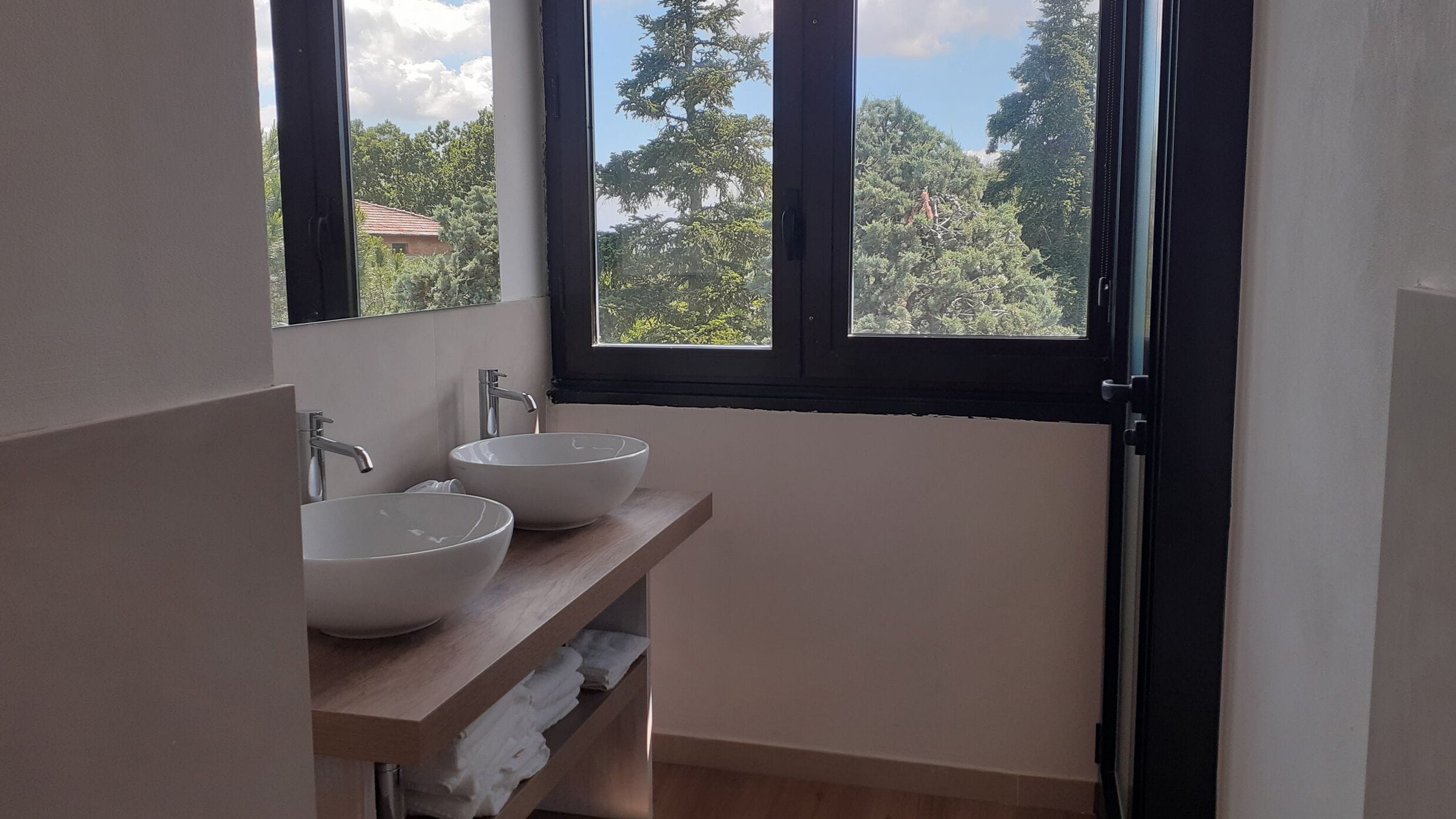 A bathroom with two white ceramic sinks, a wooden shelf, and a window with a view of trees and a building.