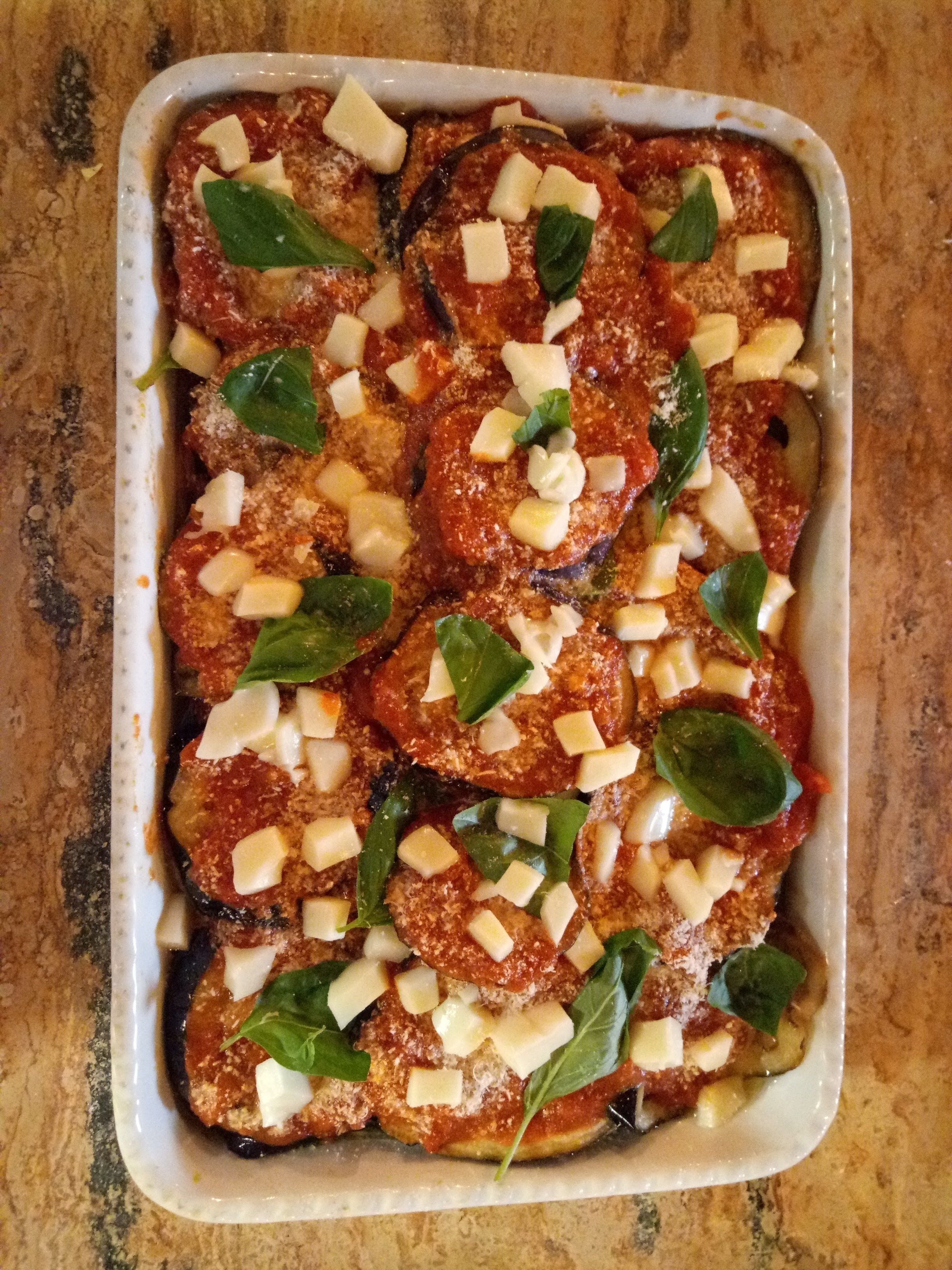 Una fuente rectangular con berenjena en rodajas cubierta de queso y albahaca, sobre una superficie texturizada.