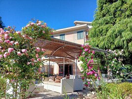 A covered outdoor seating area with a sofa and chairs, surrounded by flowering plants and a pergola.