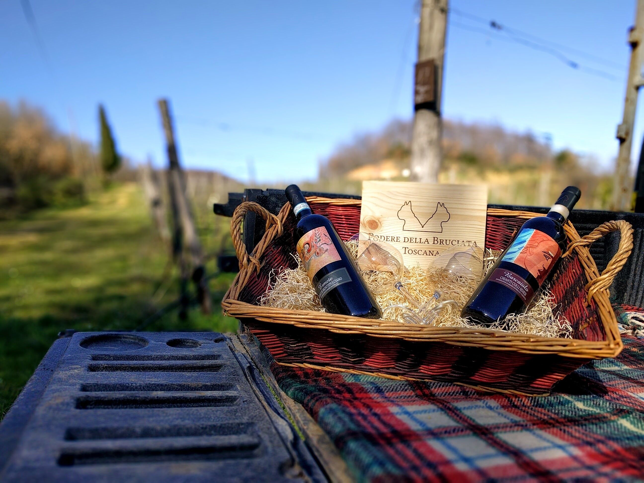 Una cesta de mimbre con dos botellas de vino y una etiqueta que dice “Fodere della Bruciata Toscana” sobre una manta a cuadros.