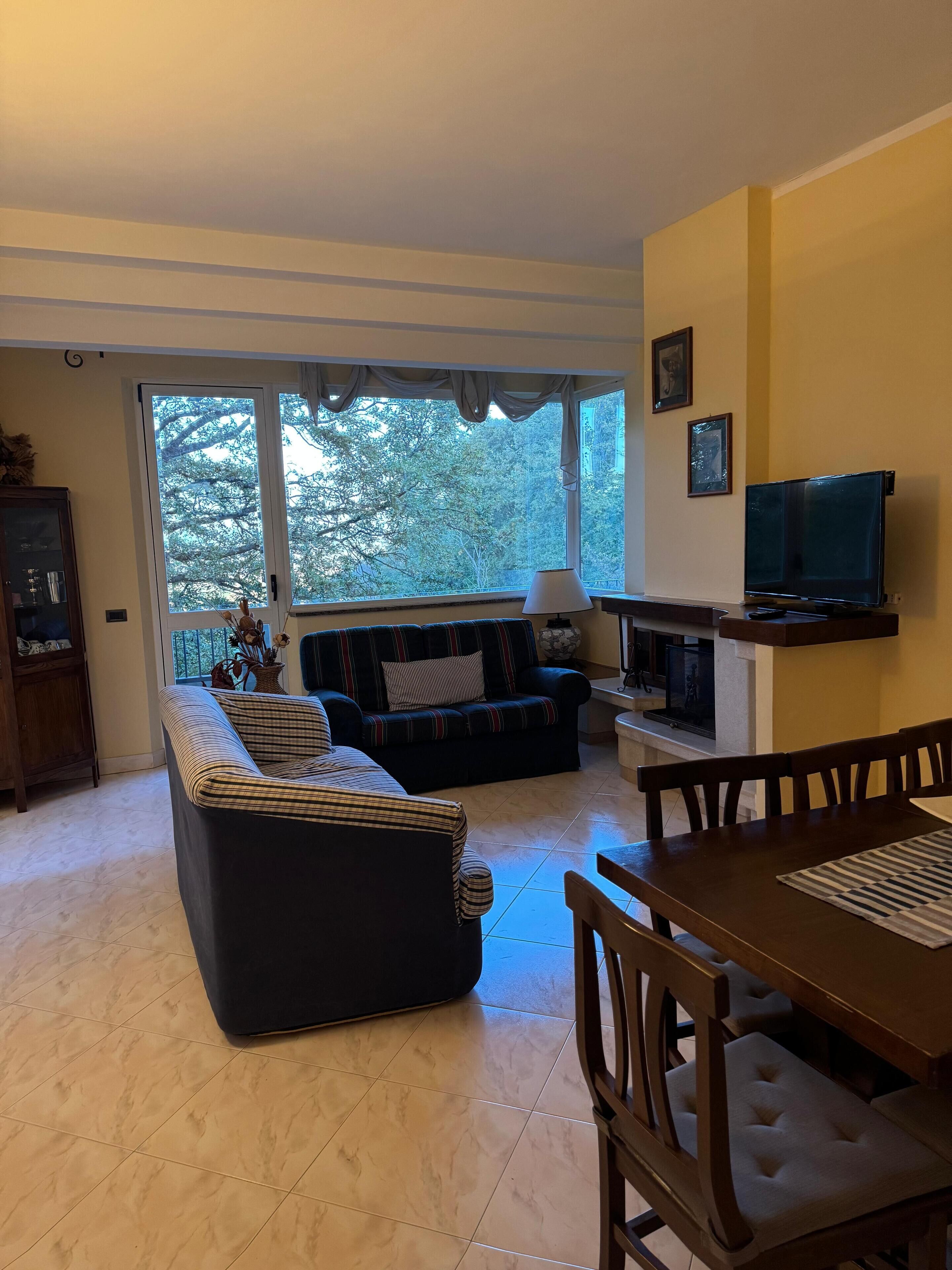Ein Wohnzimmer mit einer Couch, einem Fernseher, einem Esstisch und einem Fenster mit Blick auf Bäume.