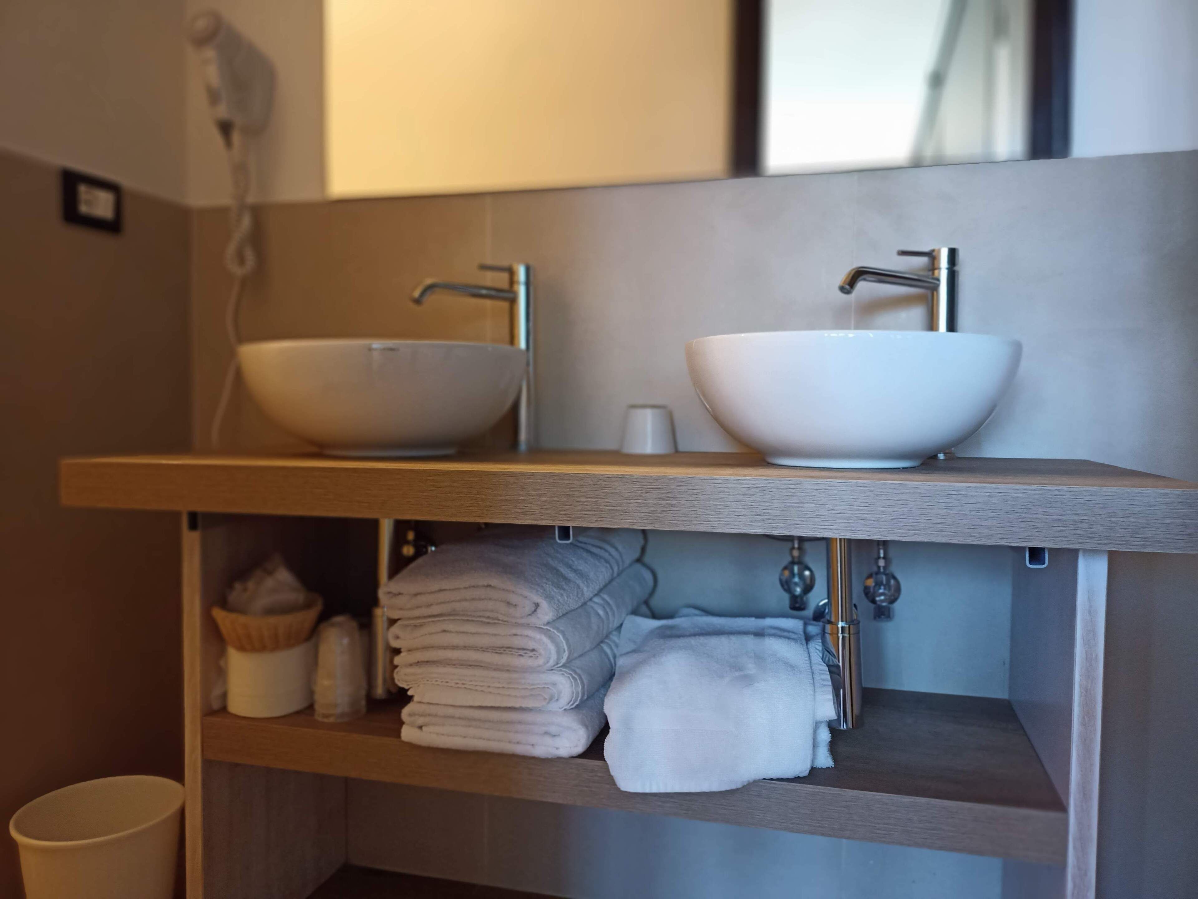 A modern bathroom with two white bowls on a wooden shelf, a towel rack below, and a hairdryer on the wall.