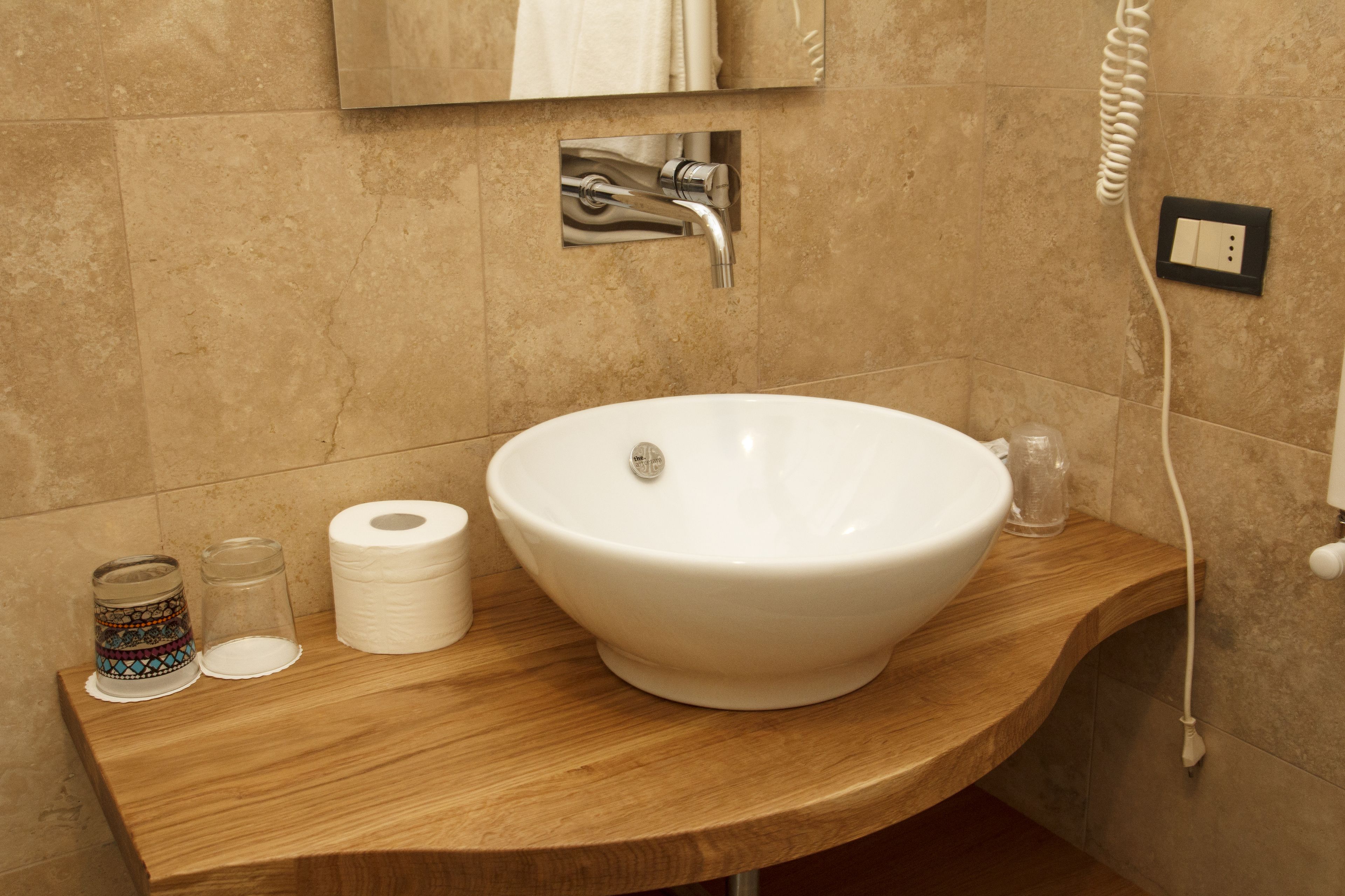 Un baño con un lavamanos grande de color blanco, un mueble de tocador de madera, un espejo, un dispensador de toallas y un teléfono fijo en la pared.