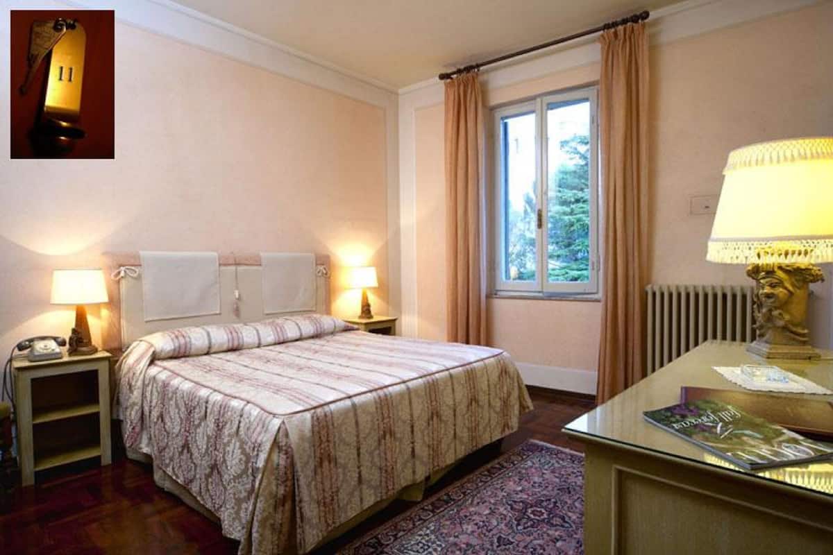 Habitació d'hotel amb un llit, dues làmpades de nit, una finestra amb cortines, un radiador, un escriptori amb una làmpada i una revista.