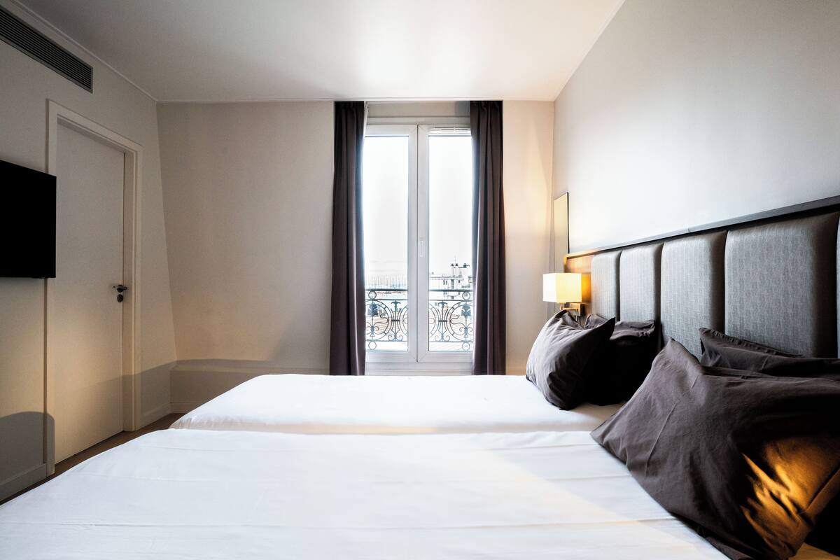 A hotel room with a large bed, a TV, a window with curtains, and a balcony view.