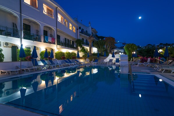 A swimming pool with lounge chairs and umbrellas, a building with balconies, and a night sky with a visible moon.