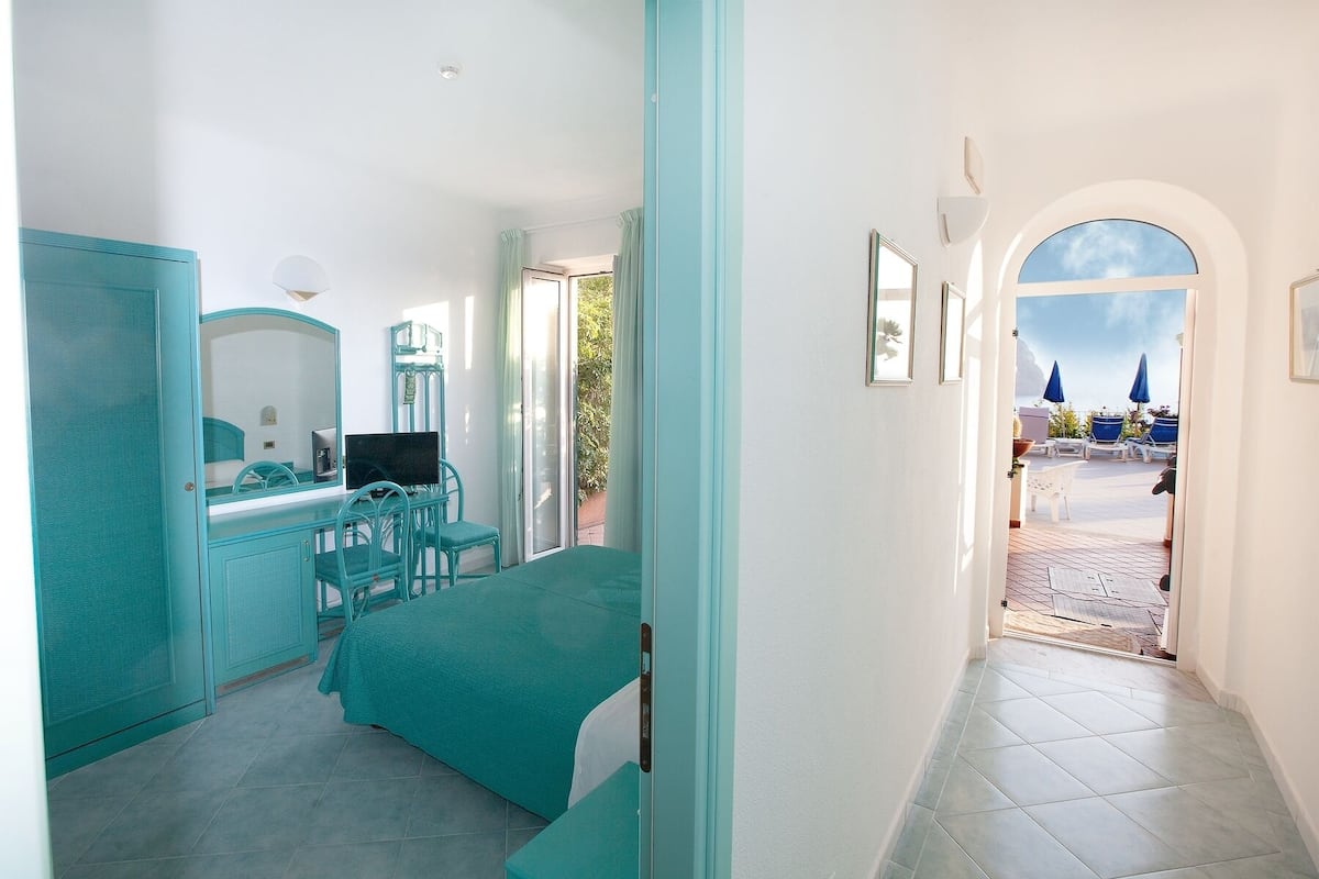 A hotel room with a bed, a desk with a computer, a chair, and a window. There is a hallway leading to an outdoor area with umbrellas and a view of the sea.