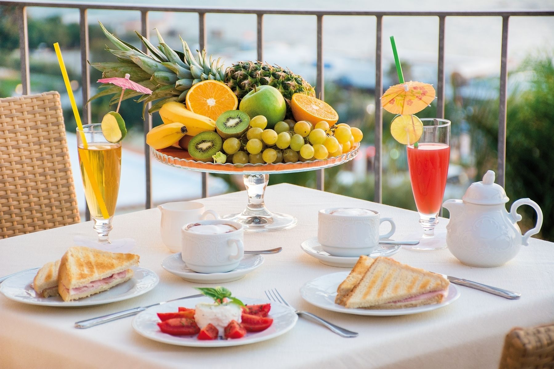 A table set with a fruit bowl, a glass of juice, two cups of tea, a teapot, and sandwiches.