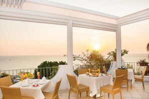 A terrace with tables set for breakfast, overlooking the sea during sunset.