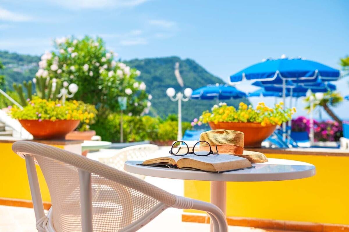 A white wicker chair, an open book, and a pair of glasses on a white table with a yellow wall in the background.
