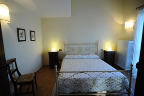 Un dormitorio con estructura de cama metálica, dos mesitas de noche con lámparas, una silla y un radiador.