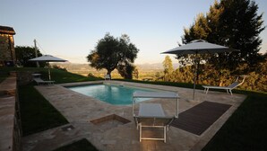 Una piscina con sillas de descanso y sombrillas, rodeada por un patio de piedra y vistas al campo.