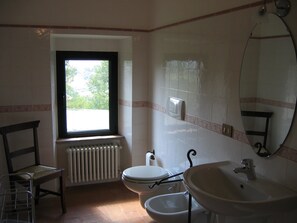 Un baño con inodoro, bidé, lavamanos, espejo, radiador, ventana y una silla.