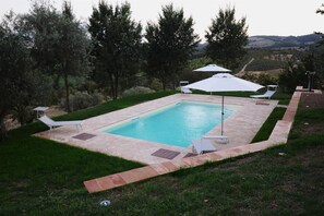 Una piscina rectangular con solárium de baldosas, rodeada por un bajo muro de piedra y césped. Hay dos sombrillas blancas y dos sillas reclinables.