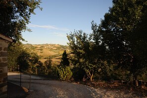 Paisaje rural con una edificación de piedra, un camino de grava y vistas a colinas onduladas y árboles.