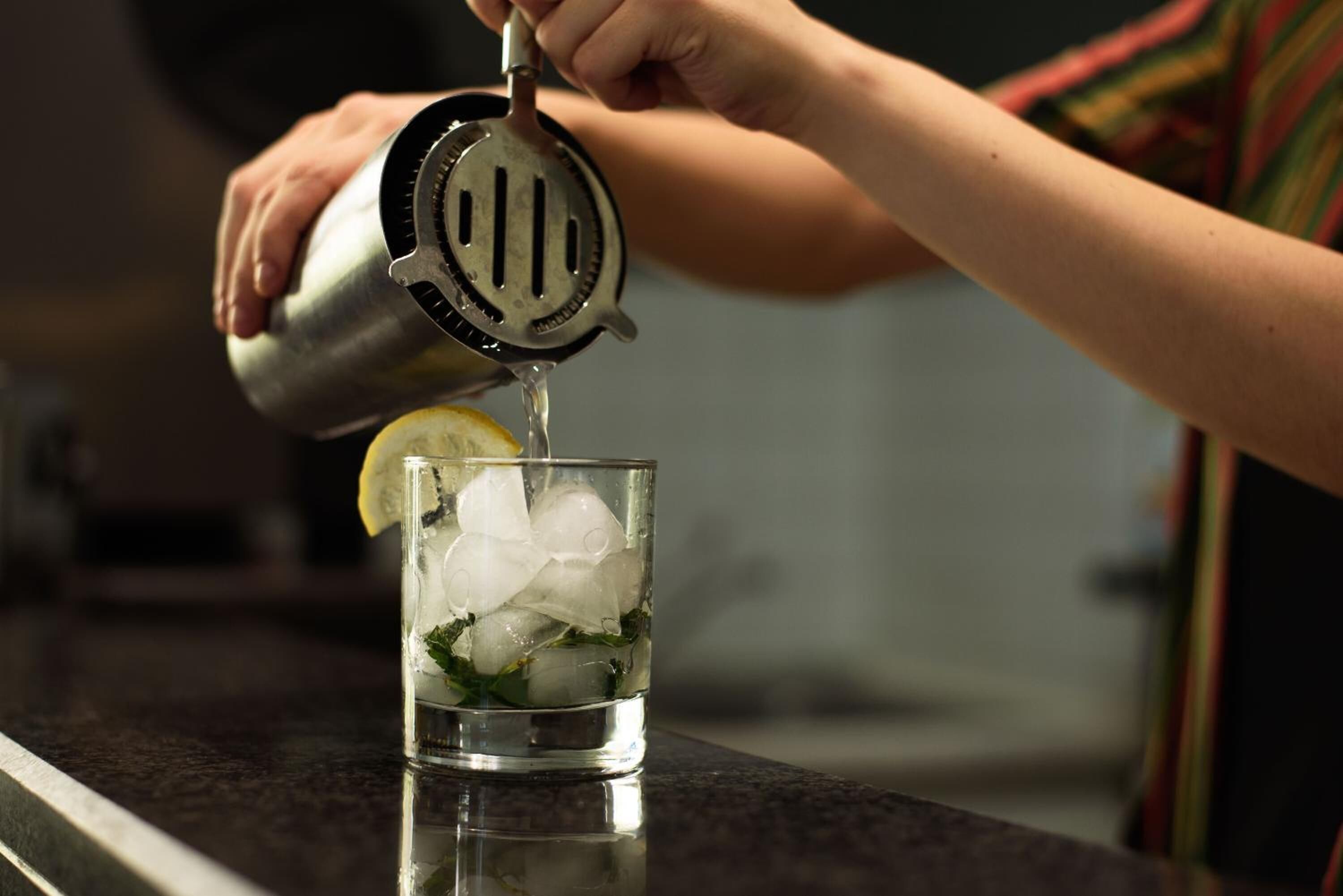 A person pouring a drink from a metal shaker into a glass with ice and a lemon slice.