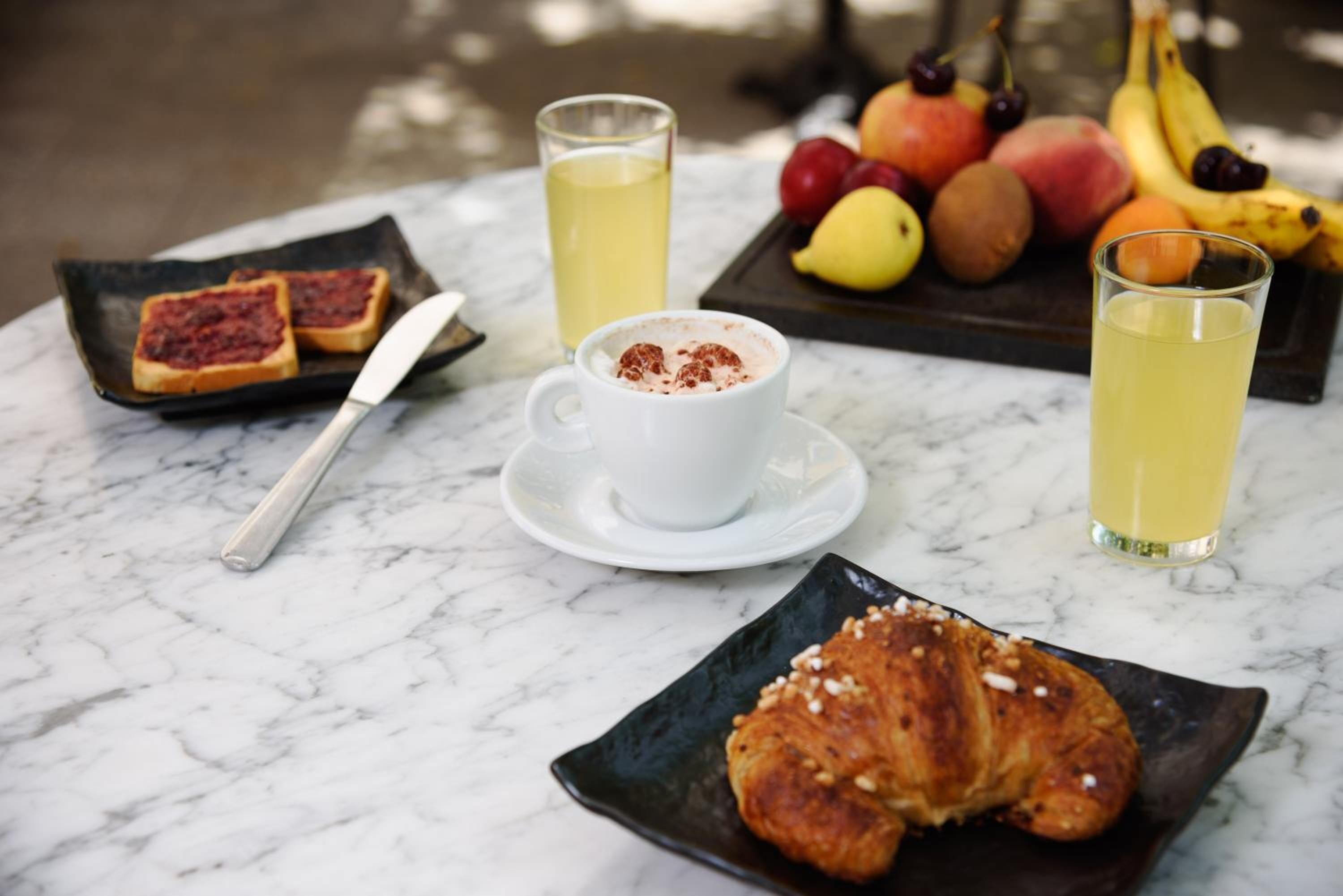Un desayuno con tostadas, un croissant, una taza de café y un plato de fruta sobre una mesa de mármol.