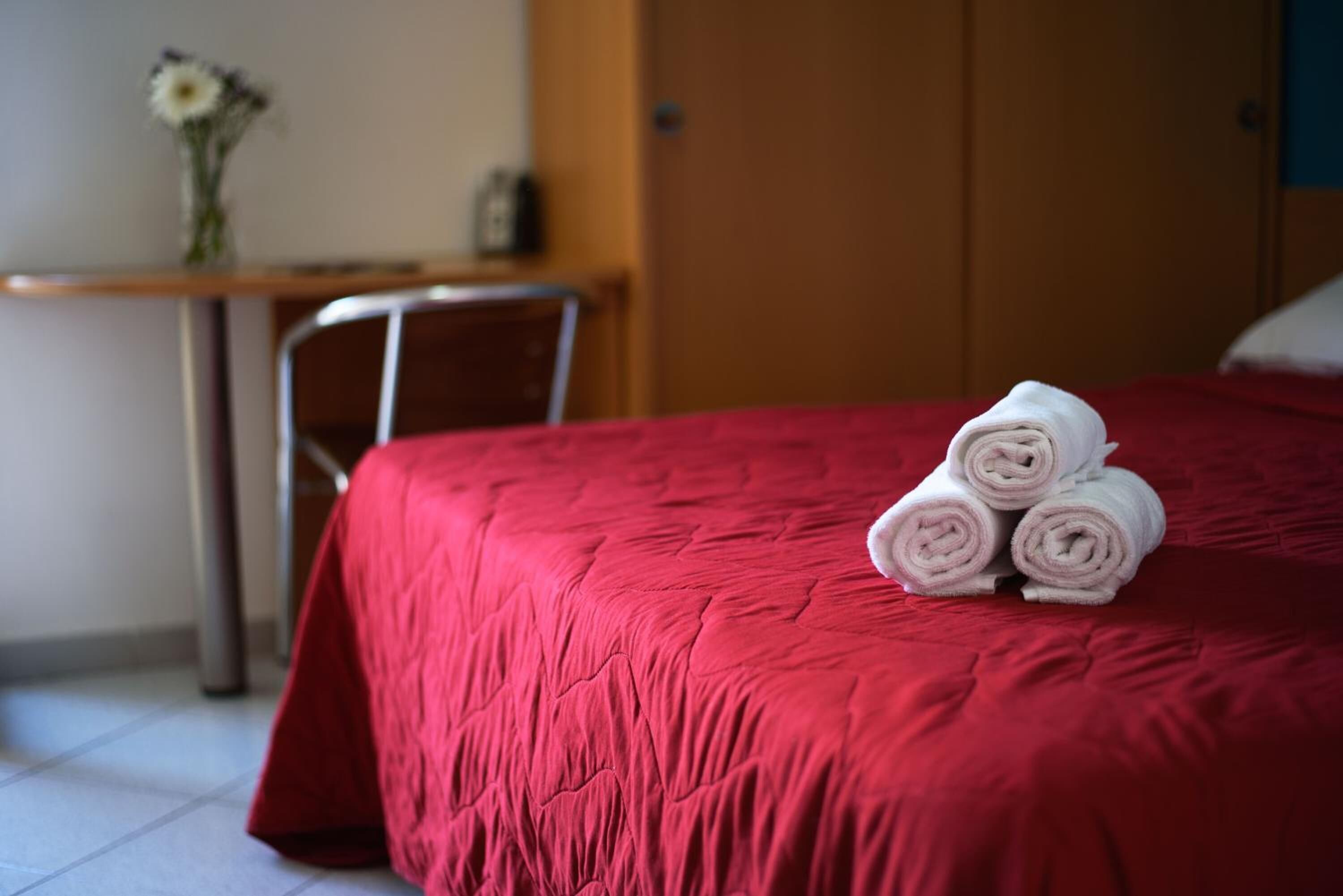 A hotel room with a red bedspread, a neatly made bed with rolled towels, a wooden desk with a vase of flowers, and a chair.