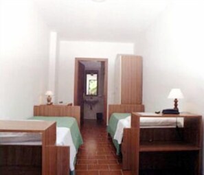 Una habitación con dos camas individuales, un armario de madera, un escritorio con una lámpara y una puerta que da a otra habitación.