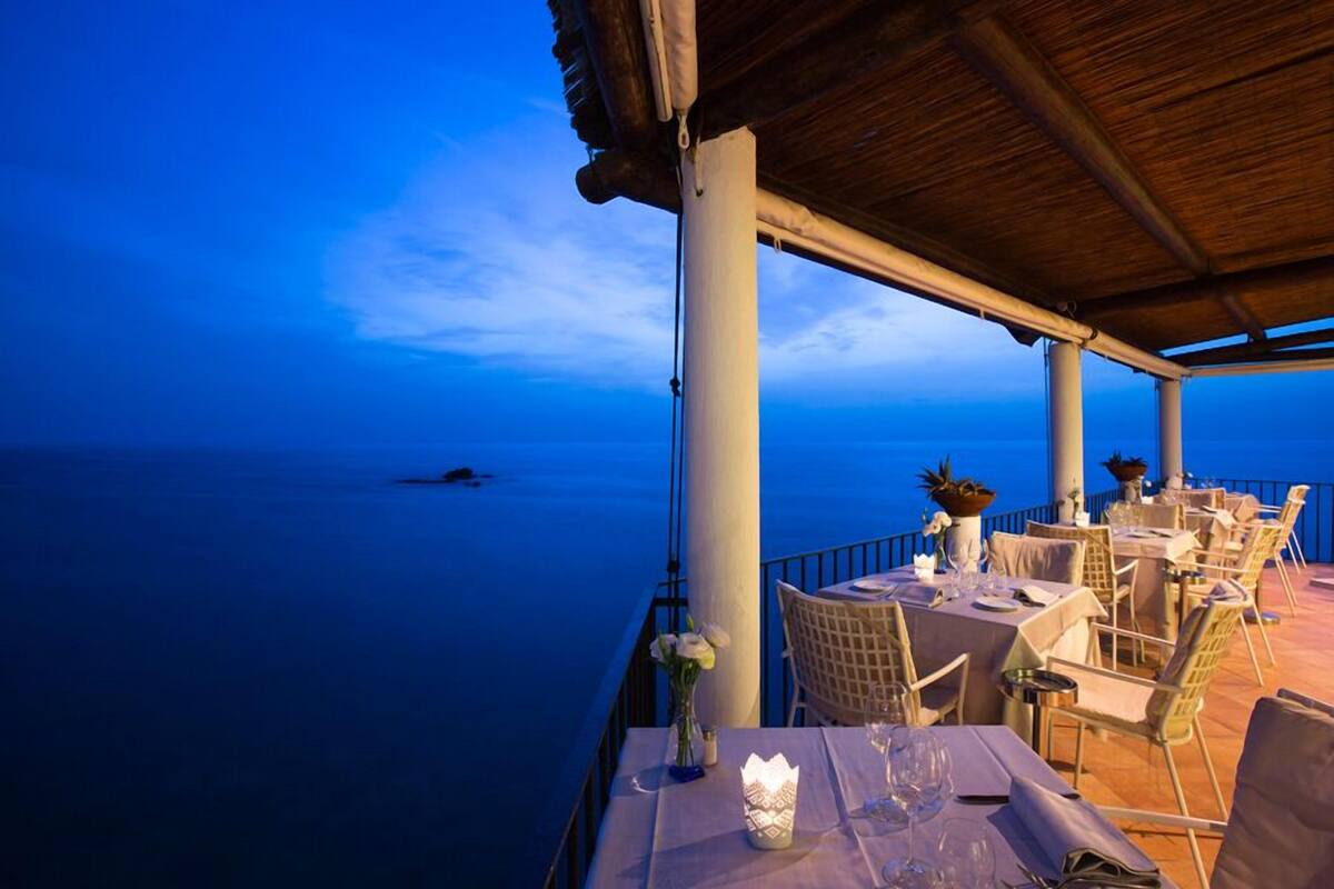 Un área de comedor al aire libre con mesas dispuestas para dos, con vista a un mar tranquilo.