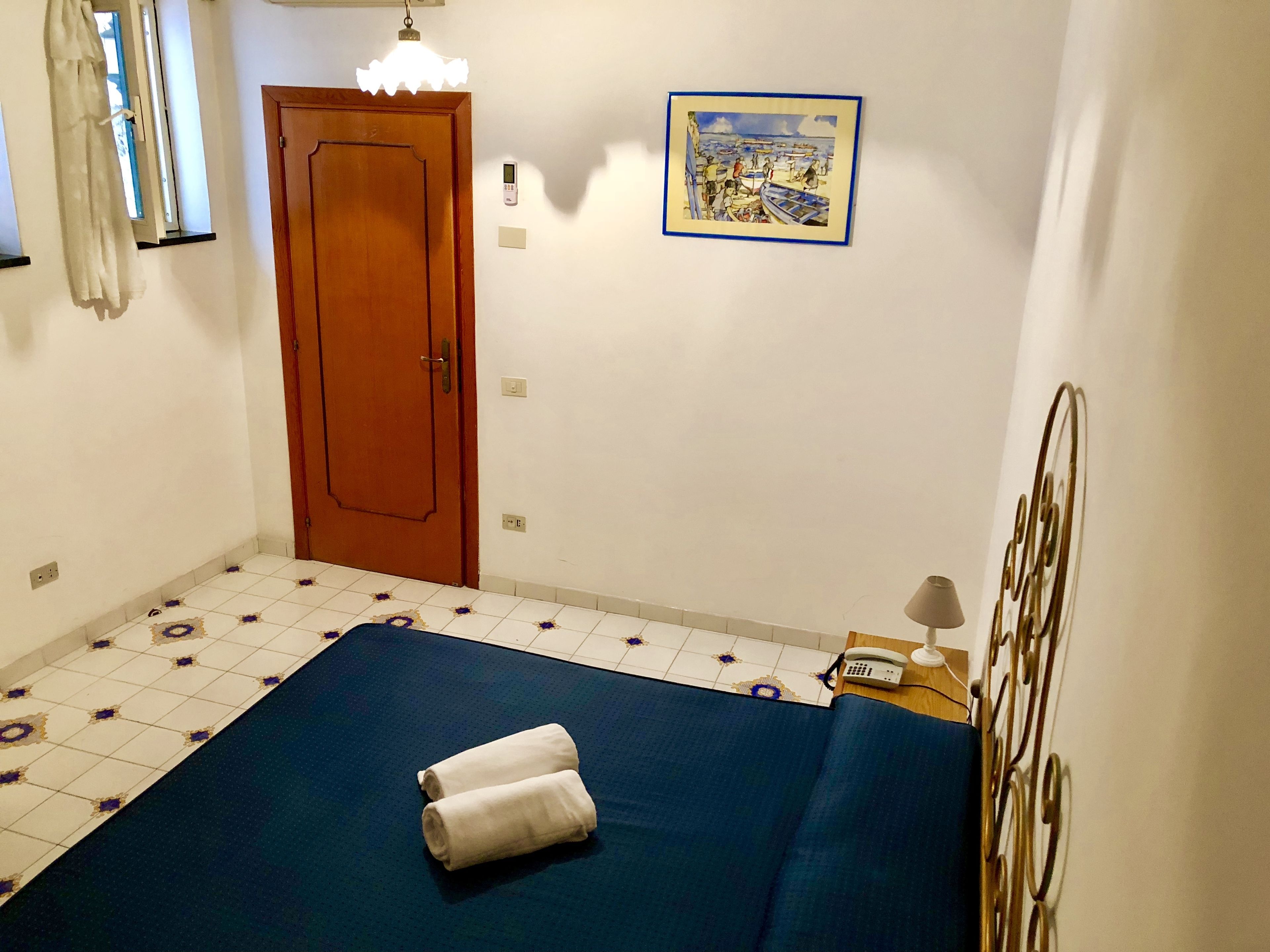 A room with a bed, a wooden door, a wall-mounted light, a painting, a telephone, and a decorative metal piece.