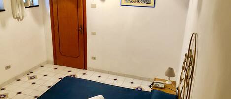 A room with a bed, a wooden door, a wall-mounted light, a painting, a telephone, and a decorative metal piece.