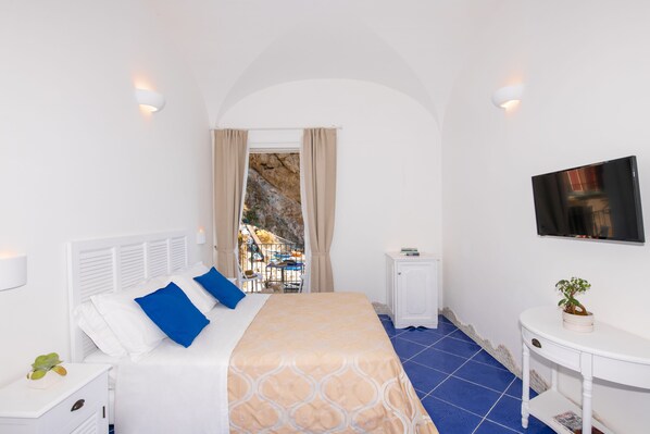 Une chambre d’hôtel avec un lit, une télévision, une petite table avec une plante et une vue sur un balcon aménagé avec du mobilier d’extérieur.