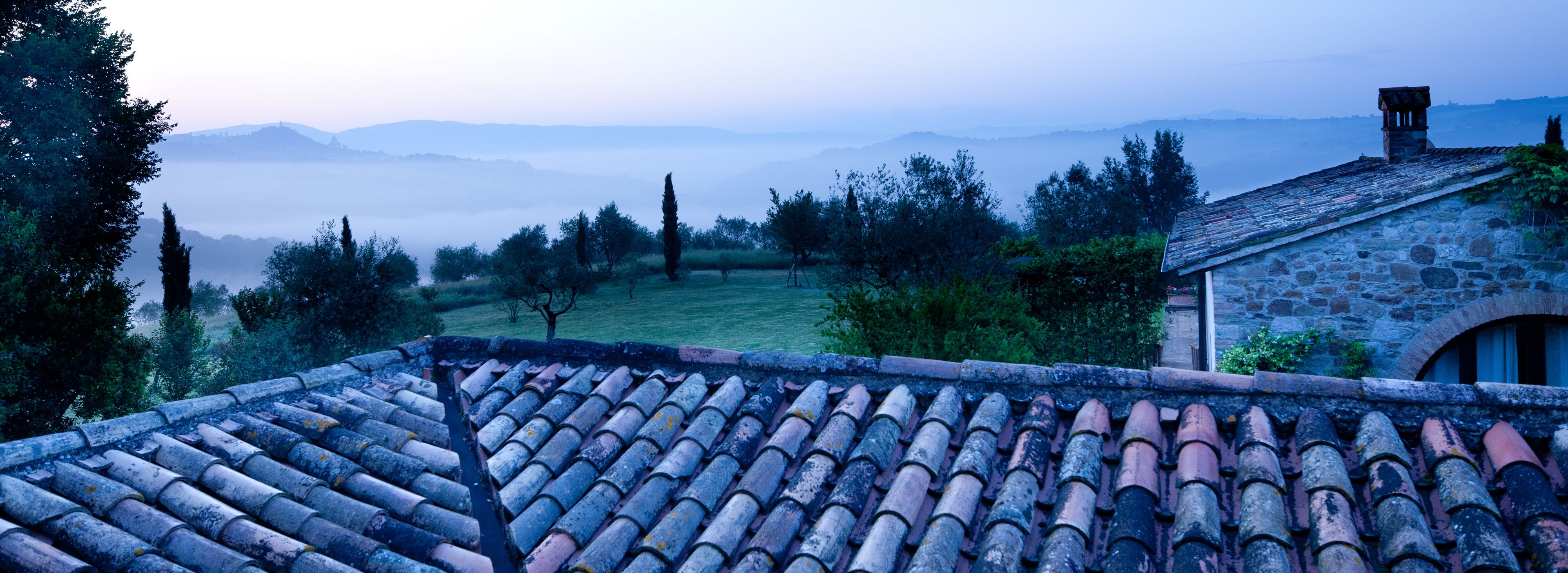 A stone building with a tiled roof overlooking a misty valley with trees and distant mountains.