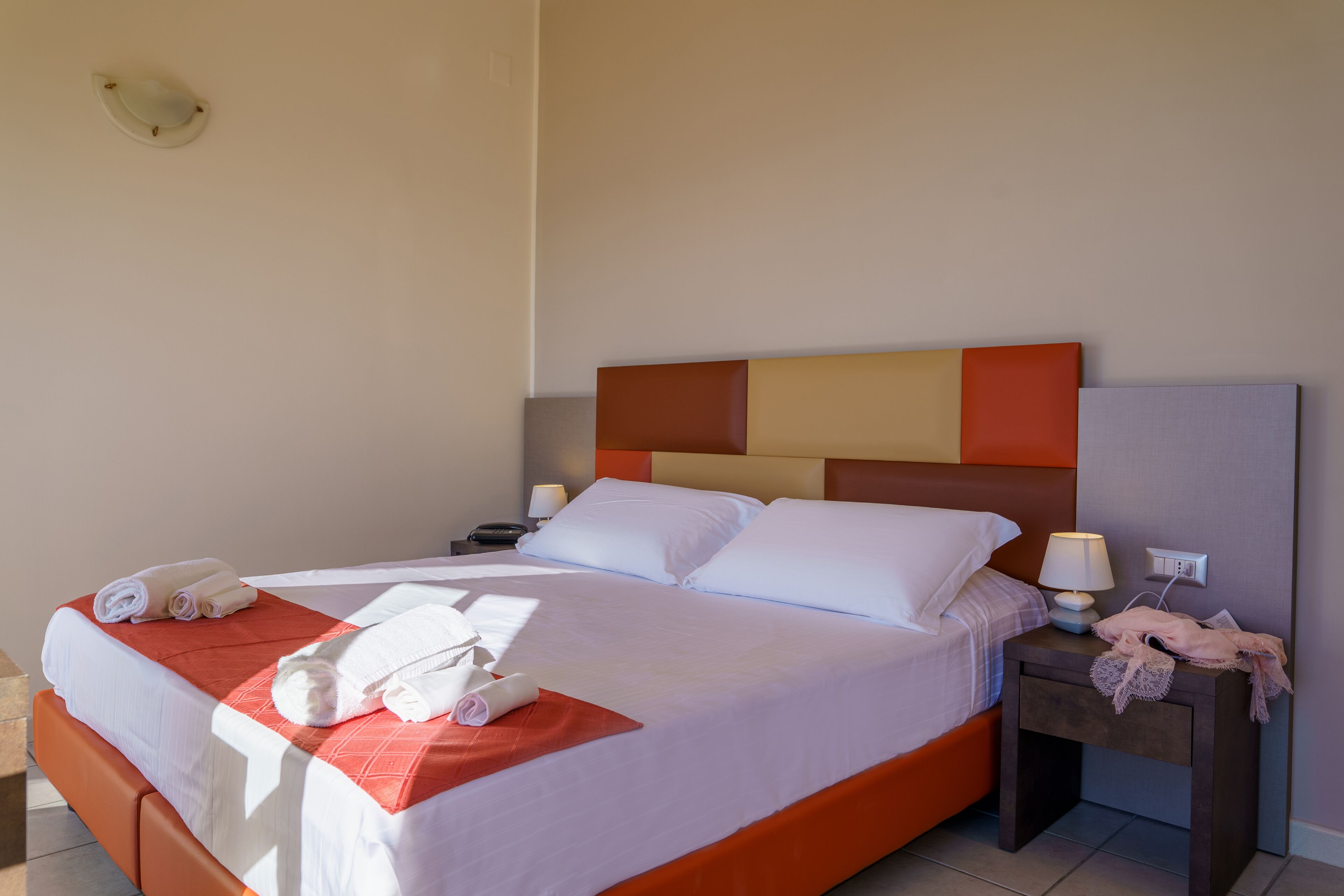 Una cama bien hecha con sábanas blancas y un detalle rojo, una mesita de noche con una lámpara, y un aplique en la pared.