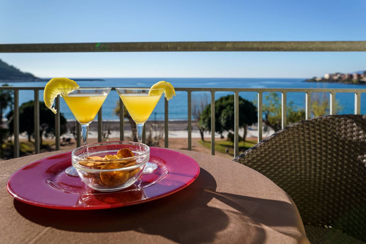 A table with two martini glasses and a bowl of food on a red plate, set on a balcony with a view of the sea.