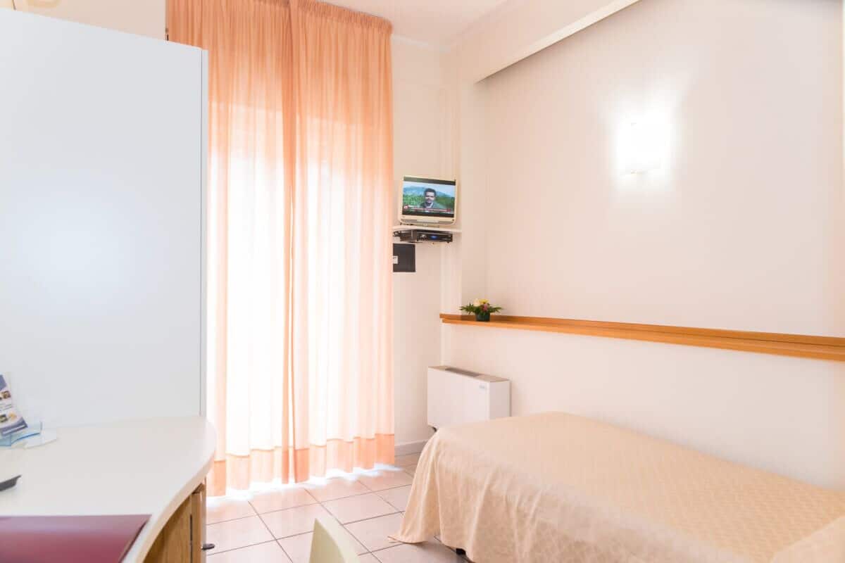 A hotel room with a bed, a desk, a television mounted on the wall, and a window with curtains.