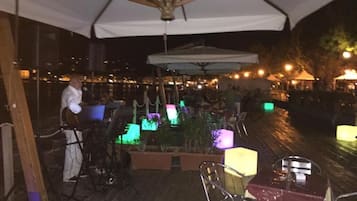 An outdoor dining area with tables and chairs, a stage with a performer, and ambient lighting.