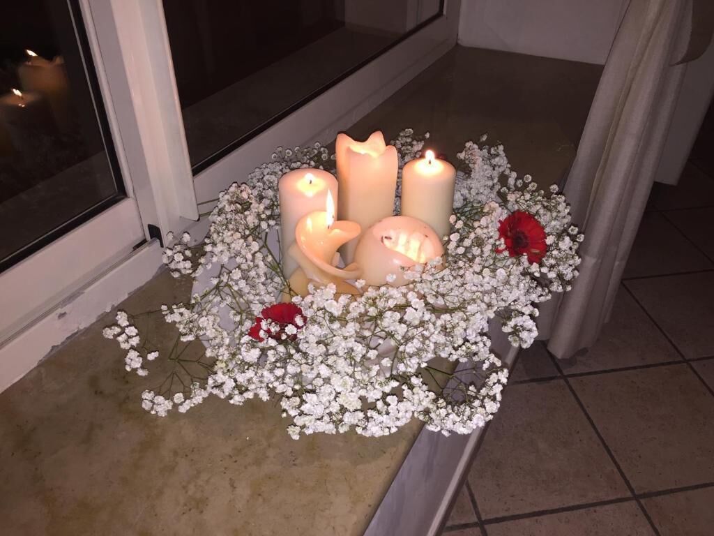 Un arreglo de velas con flores blancas y una rosa roja sobre un suelo de baldosas cerca de una ventana.