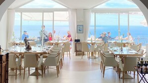 A spacious dining area with multiple tables and chairs, large windows offering a view of the sea, and a high arched ceiling. - Albergo Diffuso Bacco Furore (Furore)