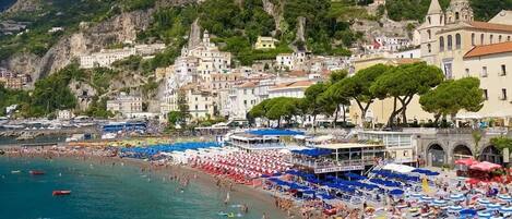 A coastal town with a beach crowded with people, a boat docked near the shore, and buildings with a mix of architectural styles.