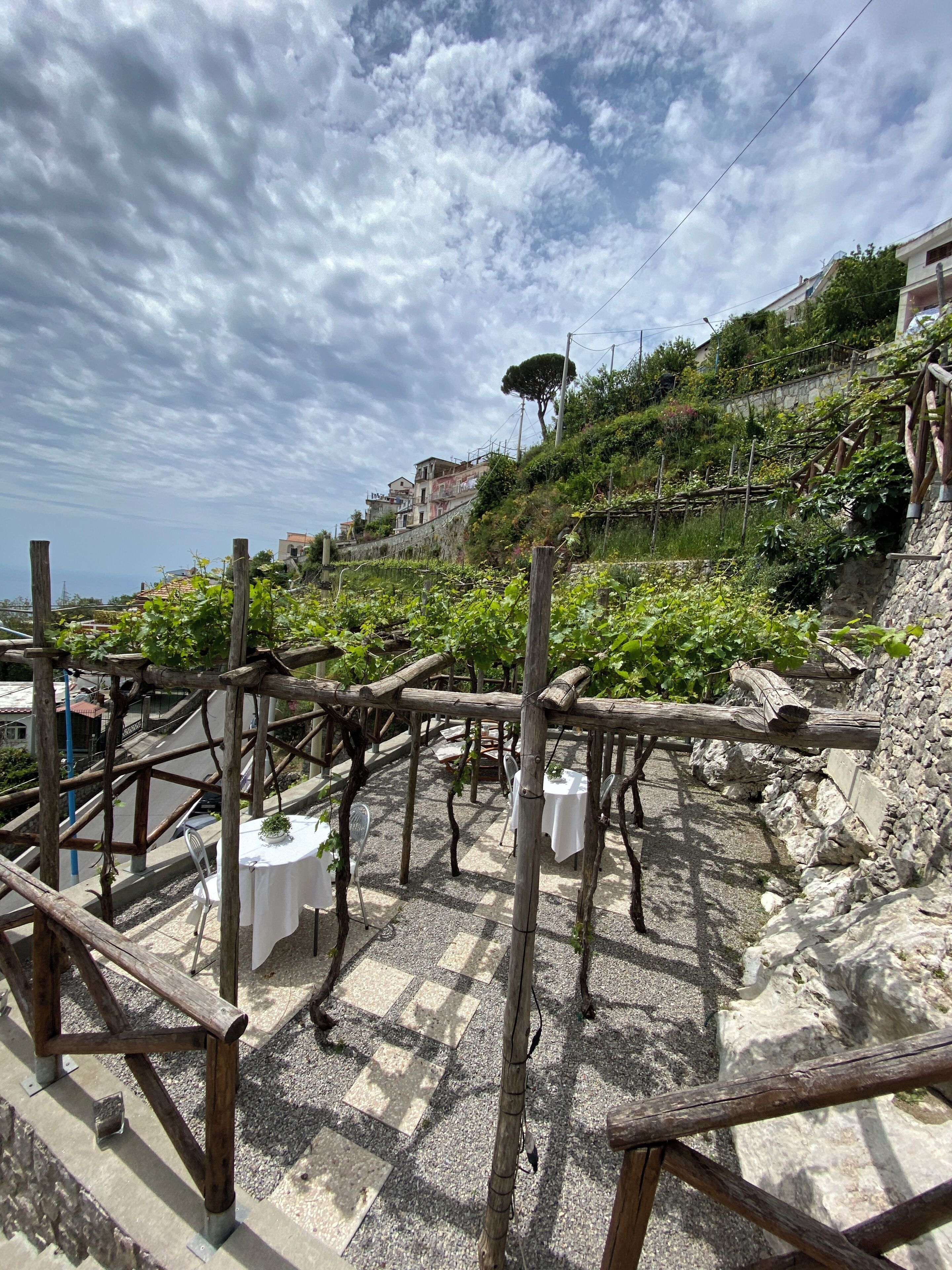 A terrace with a pergola, tables, and chairs, overlooking a hillside with buildings and greenery.