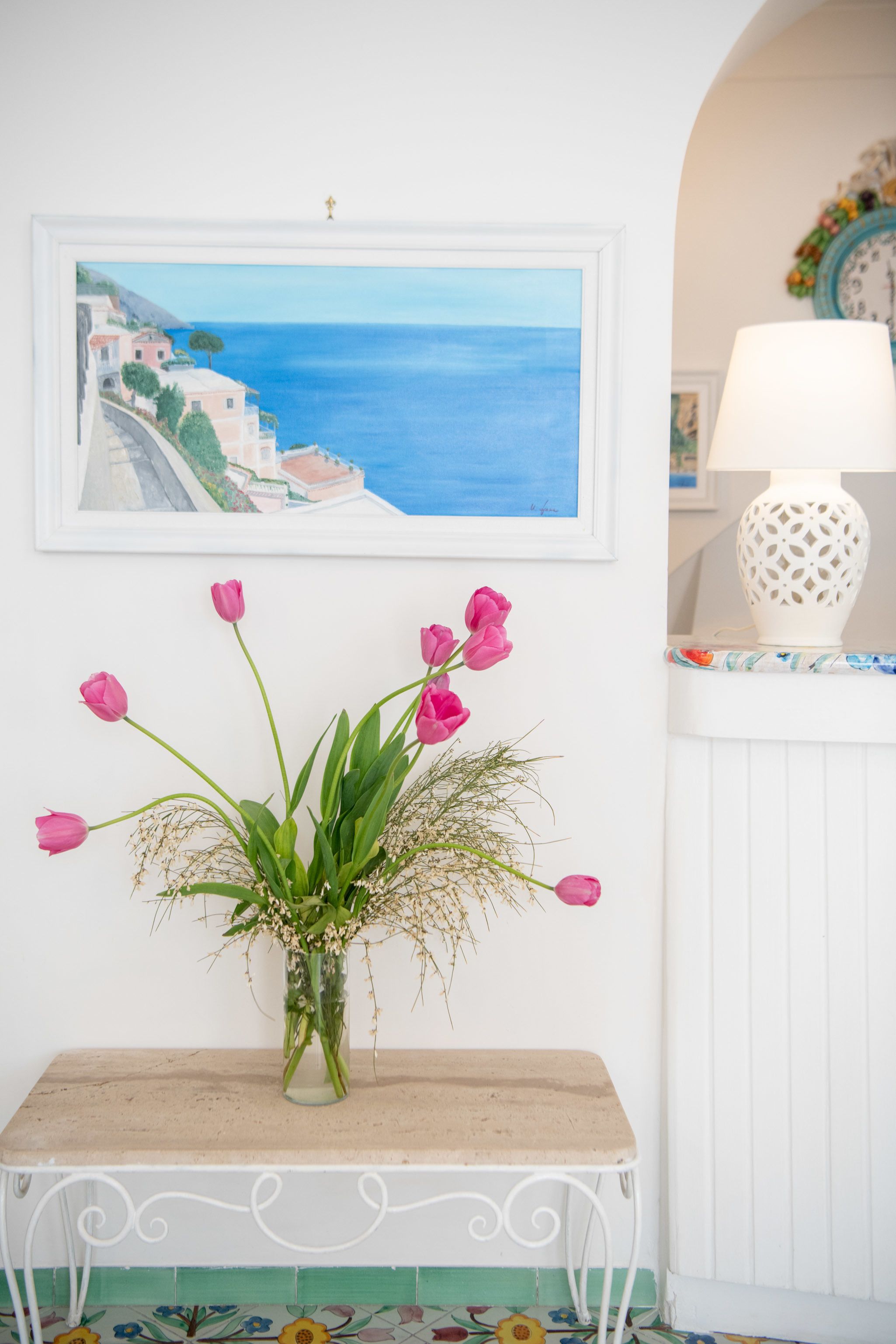 Un tableau représentant une scène côtière, encadré, un vase avec des fleurs roses et une table blanche sur laquelle se trouve une lampe.