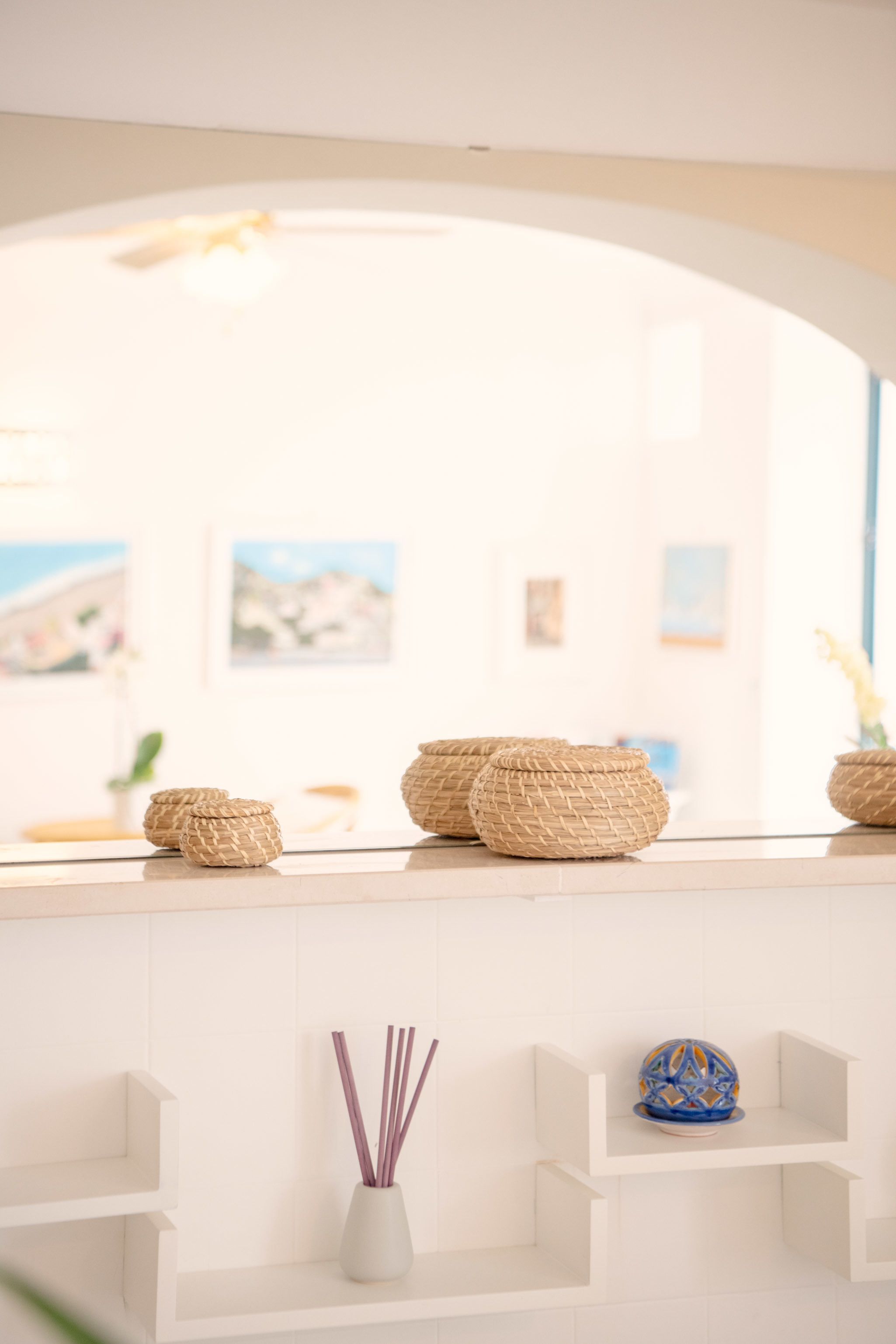 A white interior with woven baskets on a shelf, a decorative vase with sticks, and a blue patterned object on a lower shelf.