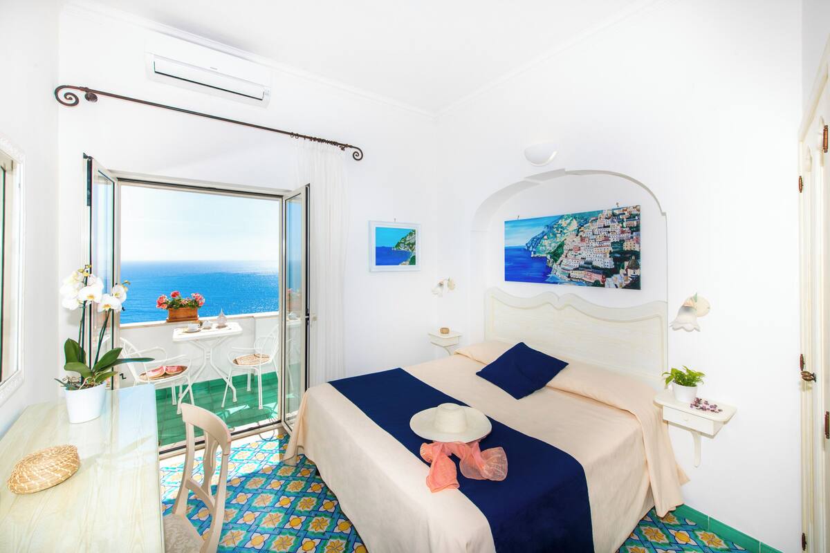 Quarto com cama, vista para o mar, varanda com cadeiras e mesa com planta.