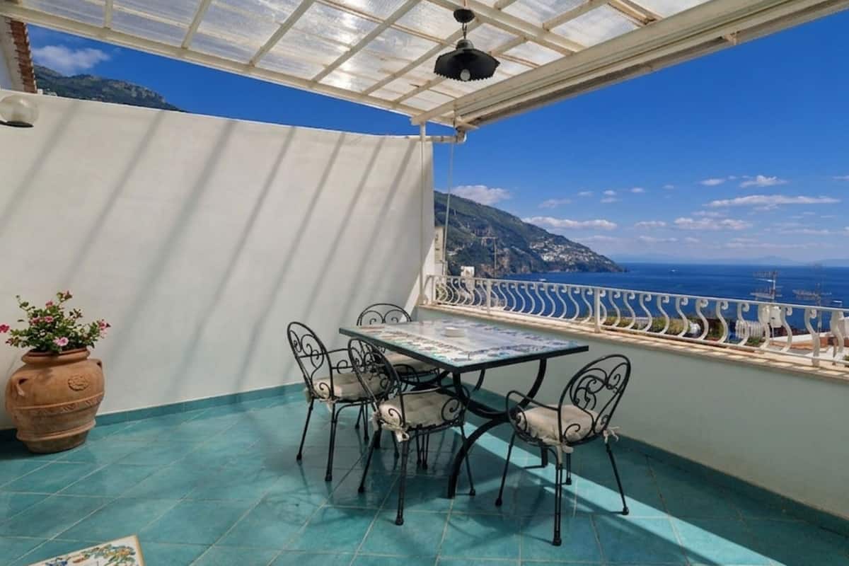 Um alpendre coberto com mesa e cadeiras, um vaso com planta, e vista para o mar e montanhas.