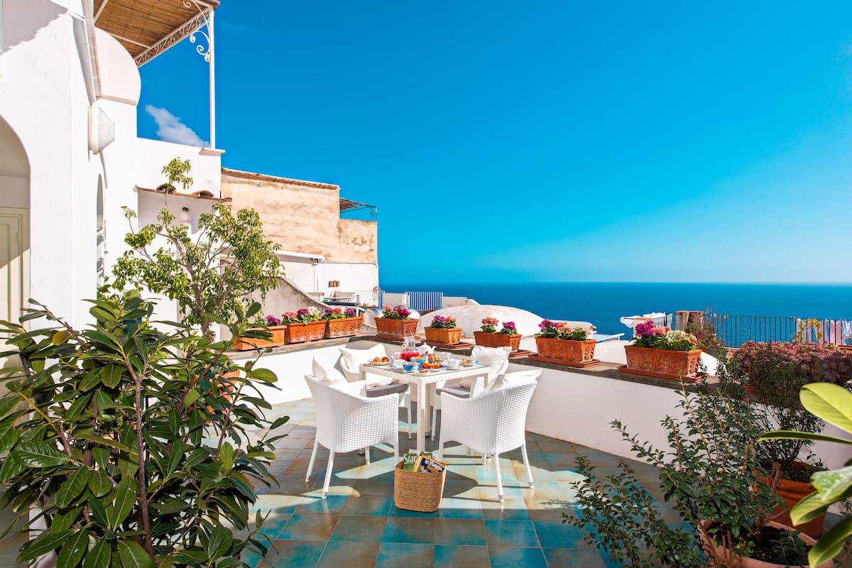 Um terraço com mobiliário de vime branco, vasos de plantas e vista para o mar.