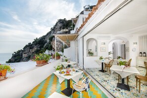 A terrace with outdoor dining tables and chairs, potted plants, and a view of the sea and cliffs. - Casa Guadagno (Positano)