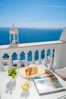 Un balcón con vista al mar, una mesa con una planta, un sombrero de paja, un vaso de bebida y una revista.
