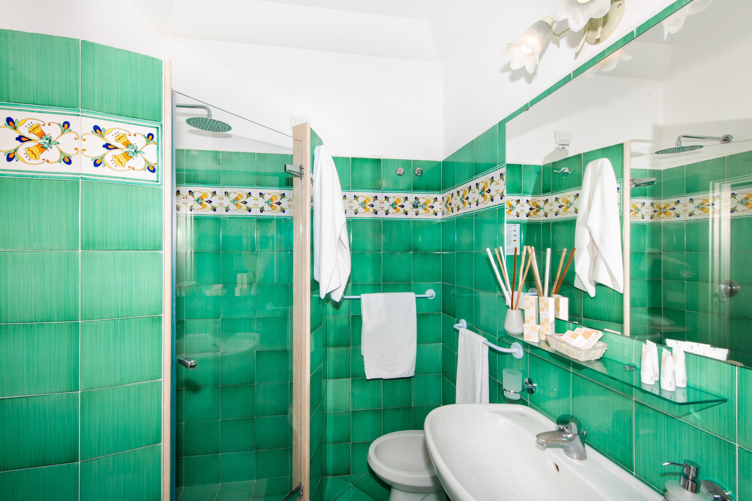 Une salle de bain avec des carreaux verts, un lavabo blanc, des toilettes, une douche en verre et un miroir.