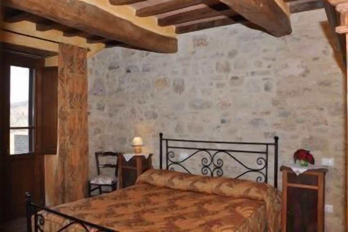 Une chambre avec un mur en pierre, des poutres en bois, un lit recouvert d’une couvre-lit à motifs, une table de chevet avec une lampe et une petite table sur laquelle est posé un vase.
