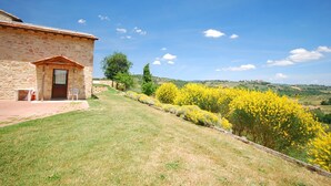 Un bâtiment en pierre avec une porte et des fenêtres, une zone herbeuse parsemée de fleurs jaunes et un ciel bleu clair.
