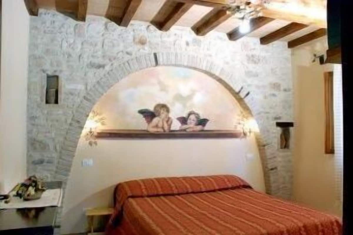 Une chambre avec un mur en pierre, une fenêtre en arc de cercle, un lit recouvert d’une couverture à rayures et une petite table avec une lampe.