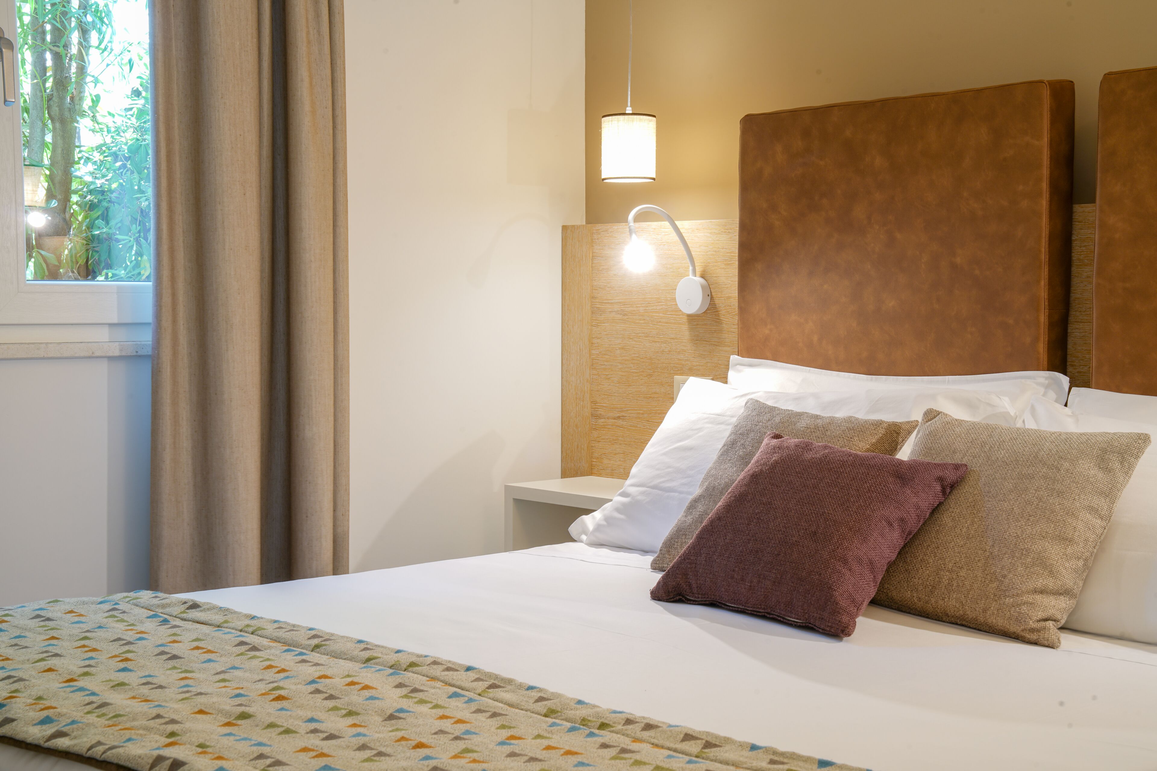 A neatly made bed with a patterned bedspread, a bedside lamp, and a view of greenery outside.