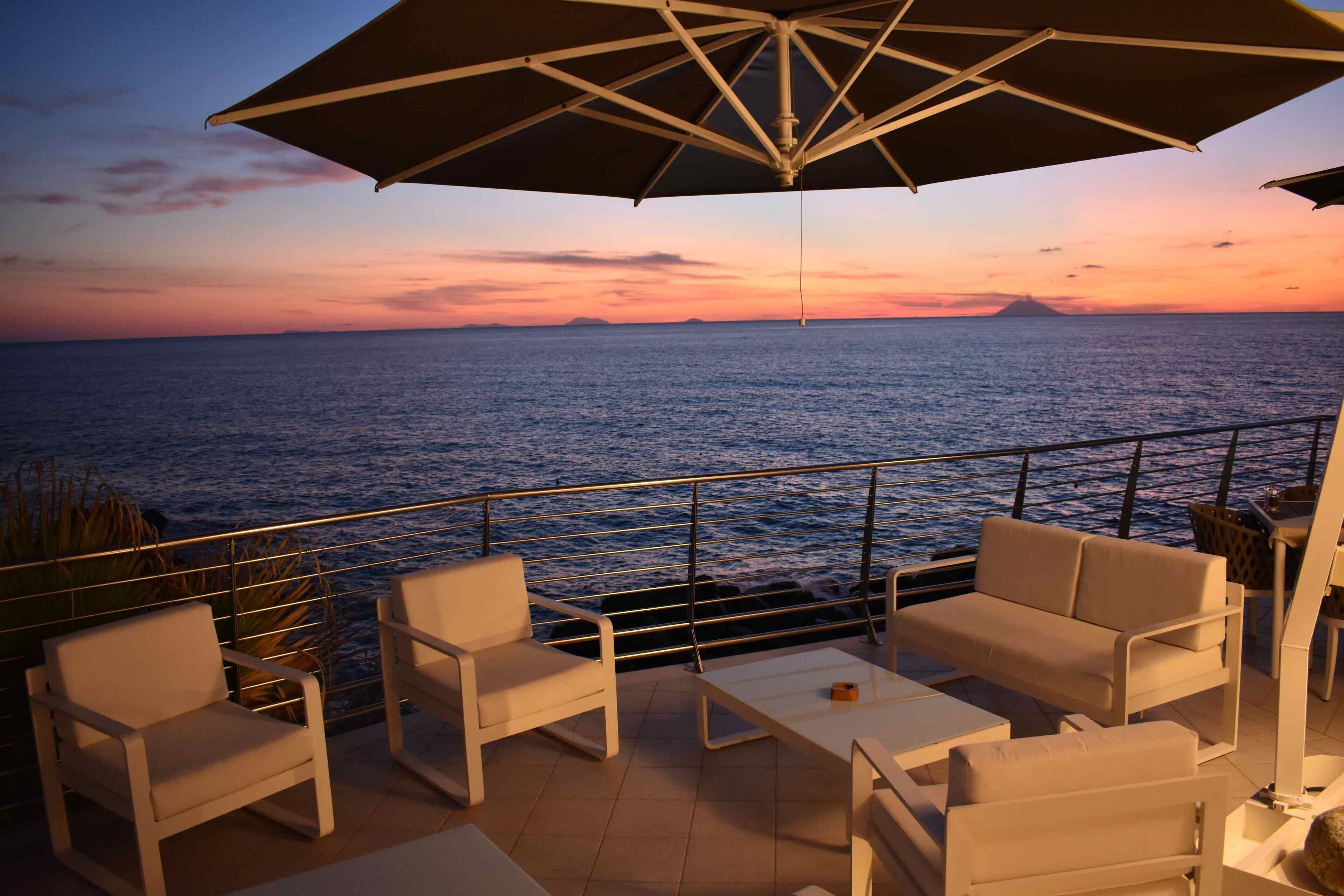 A terrace with white furniture, a large umbrella, and a view of the sea at sunset.