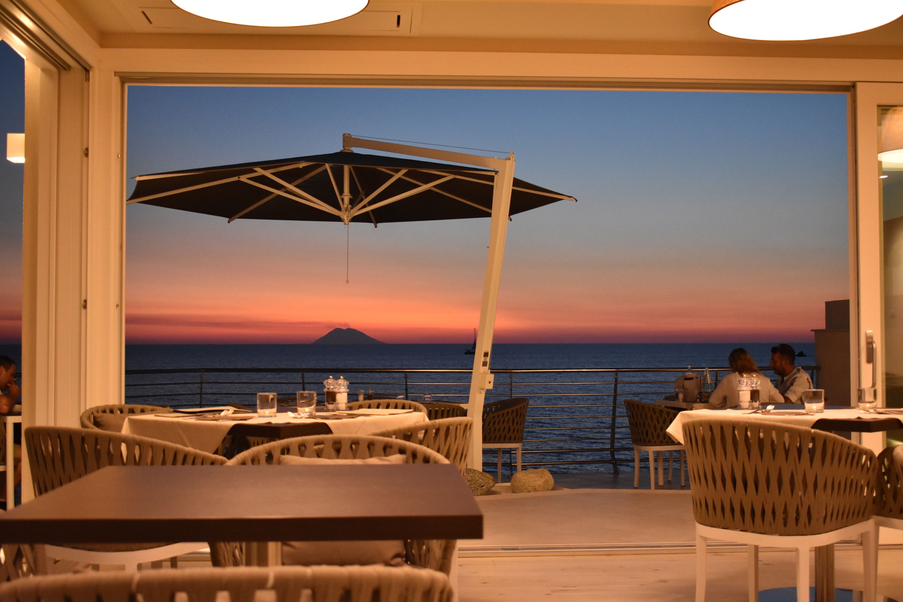An outdoor dining area with tables and chairs, a large umbrella, and a view of the ocean at sunset.